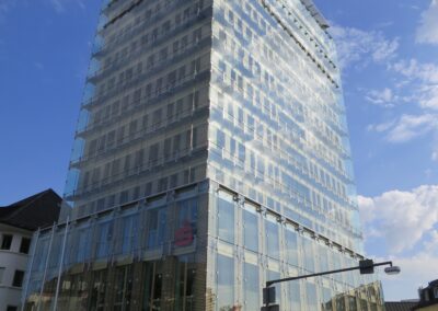 Ein modernes Hochhaus mit heller Glasfassade und regelmäßigen Fensterreihen steht an einem sonnigen Tag vor blauem Himmel.