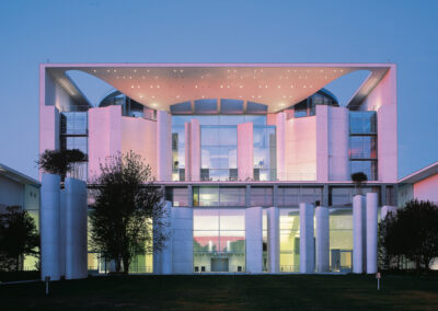 Ein modernes Gebäude mit rosa beleuchteter Fassade, großen Glasflächen und markanten weißen Säulen prägt die Architektur bei Dämmerung.