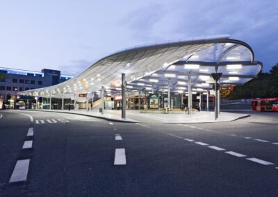 Eine moderne, geschwungene Busstation mit fließender Glasfassade und eleganter Stahlkonstruktion in der Abenddämmerung.