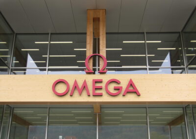 Ein modernes Gebäude mit Glasfassade und hölzernem Träger zeigt das rosafarbene Omega-Logo auf heller Fassade.