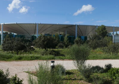 Ein modernes Gebäude mit geschwungener Membrandachstruktur und blauen Stützen steht in einer Landschaft mit mediterranen Pflanzen.