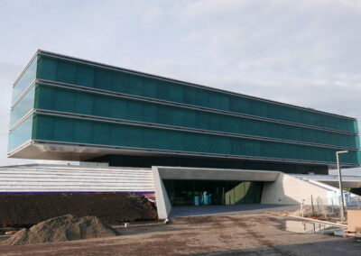 Ein modernes Gebäude mit grüner Glasfassade und horizontalen Fensterbändern schwebt über einem offenen Eingangsbereich aus Beton.