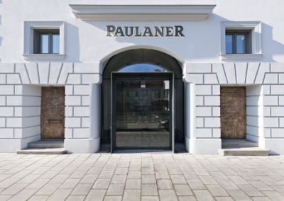 Das Bild zeigt die klassische Fassade einer Paulaner-Brauerei mit weißer Putzfläche, symmetrischen Fenstern und einem markanten gewölbten Eingangsportal.