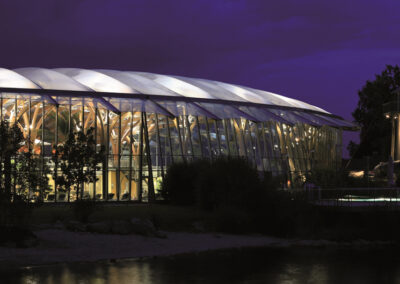 Ein modernes Gewächshaus mit gläserner Fassade wird in der Dämmerung beleuchtet und spiegelt sich im Wasser davor.