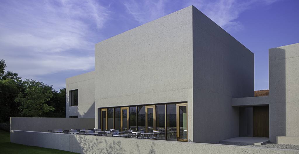 Ein modernes Betongebäude mit geometrischen Formen, großflächigen Fenstern und einer minimalistischen Architektur unter blauem Himmel.
