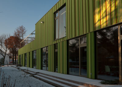 Ein modernes Gebäude mit grüner Wellblechfassade und großflächigen Fenstern steht in einer winterlichen Landschaft.