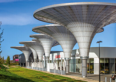 Eine moderne Architektur mit markanten, pilzförmigen Betonkonstruktionen überdacht eine Reihe von weißen Ladengeschäften unter blauem Himmel.