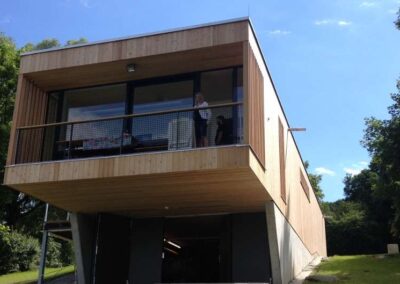 Ein modernes Wohnhaus mit Holzfassade und großem Balkon schwebt auf Stützen über einer Grünfläche.
