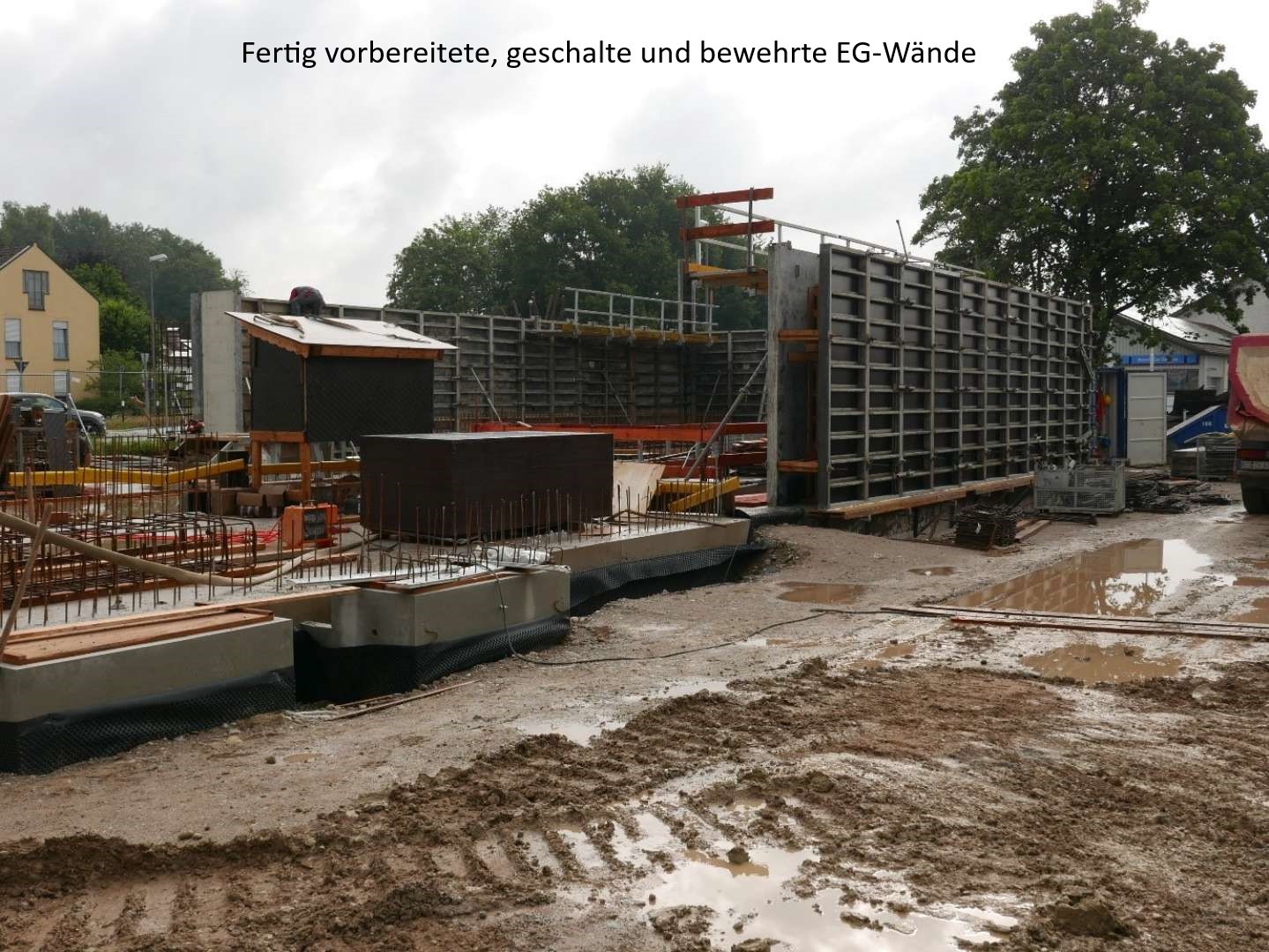 Eine Baustelle mit teilweise fertiggestellten Betonwänden, Schalungen und Bewehrungen für ein modernes Gebäude im Rohbau.