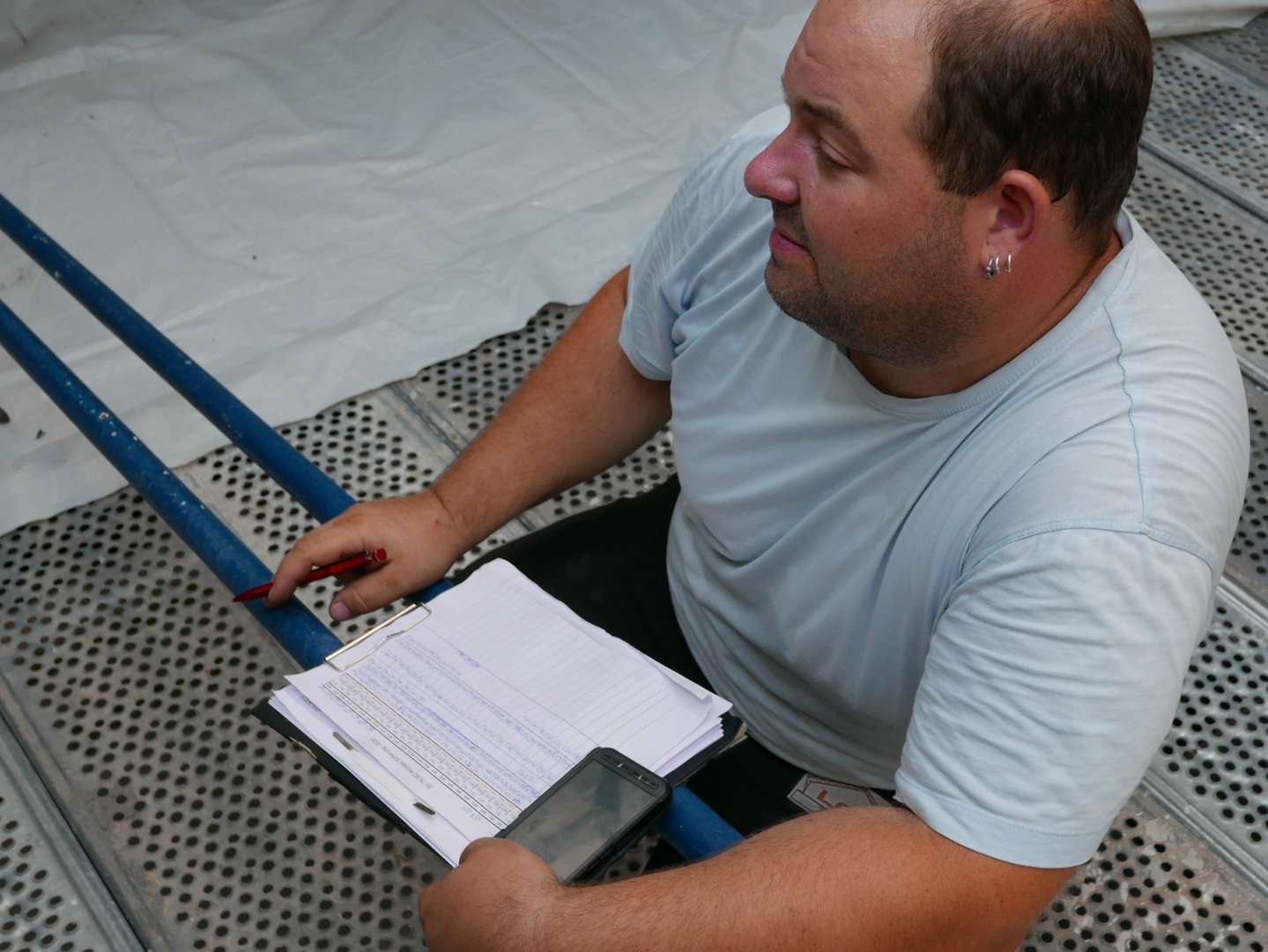 Ein Mann in hellblauem Hemd sitzt auf einem perforierten Metallgitter und prüft Baupläne mit Bleistift.