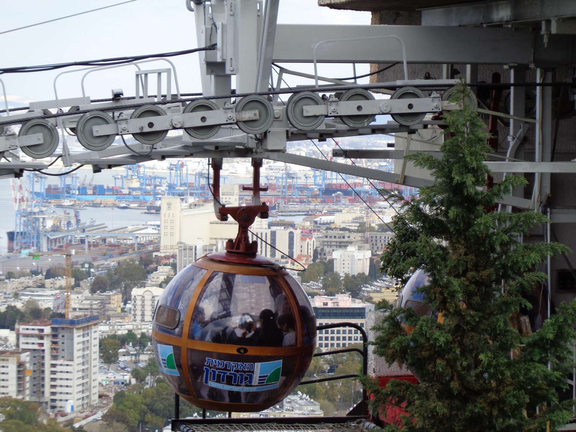 Eine Seilbahn-Gondel schwebt über einer dicht bebauten Hafenstadt mit modernen Hochhäusern und Industrieanlagen.