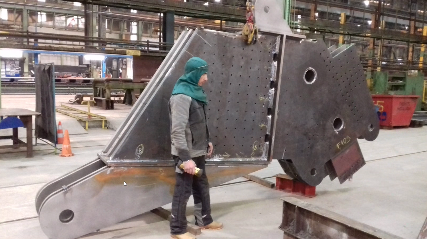 Ein Arbeiter in einer Industriehalle inspiziert große metallische Bauteile mit Lochungen und geometrischen Formen.