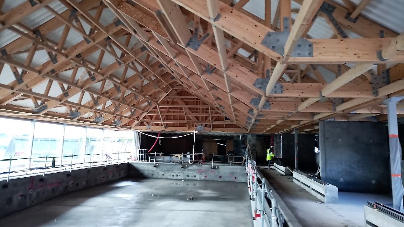 Ein moderner Rohbau mit freiliegender Holzdachkonstruktion, Betondecken und großflächiger Glasfassade unter Baugerüsten.