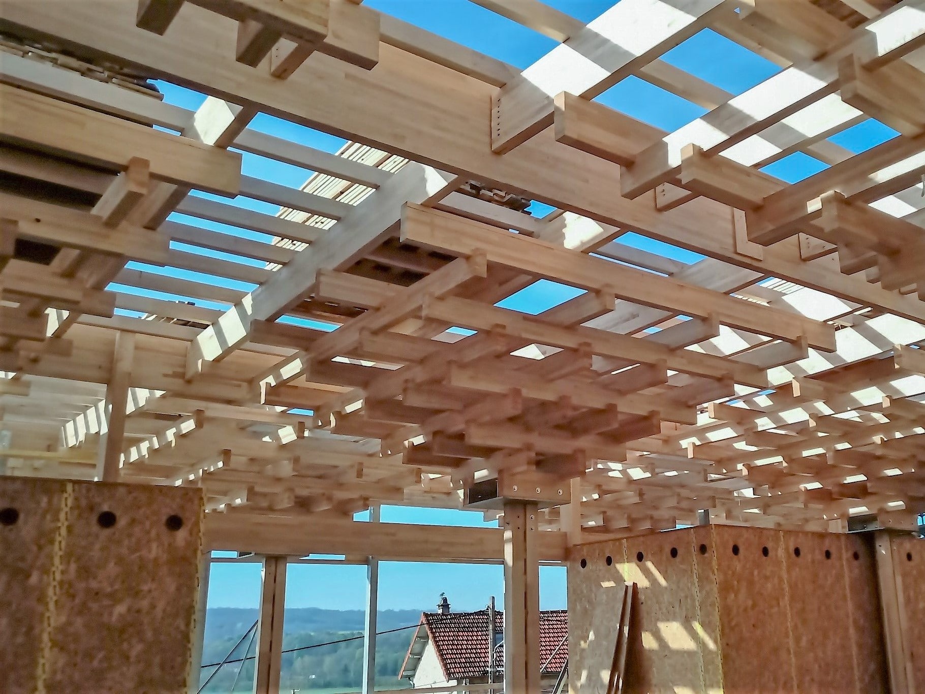 Ein moderner Holzbau mit offener Dachkonstruktion zeigt massive Balken und große Fenster mit Ausblick auf eine bergige Landschaft.