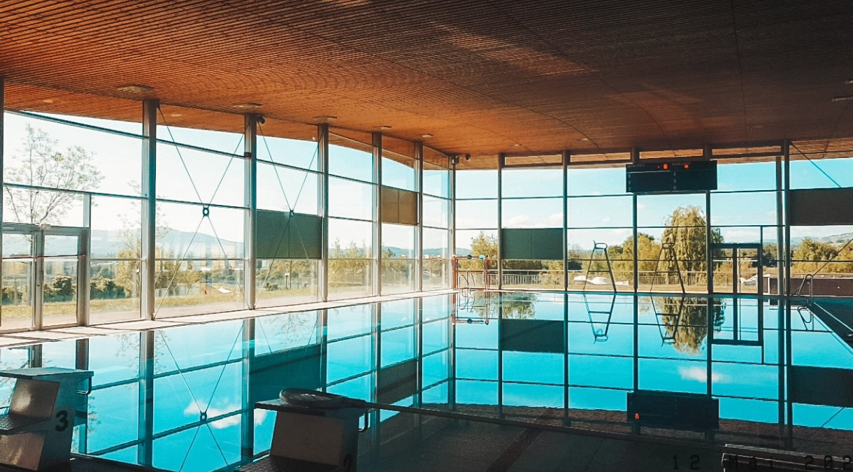 Ein modernes Hallenbad mit großflächiger Glasfassade, Holzdecke und Stahlkonstruktion bietet Blicke auf die umgebende Landschaft.