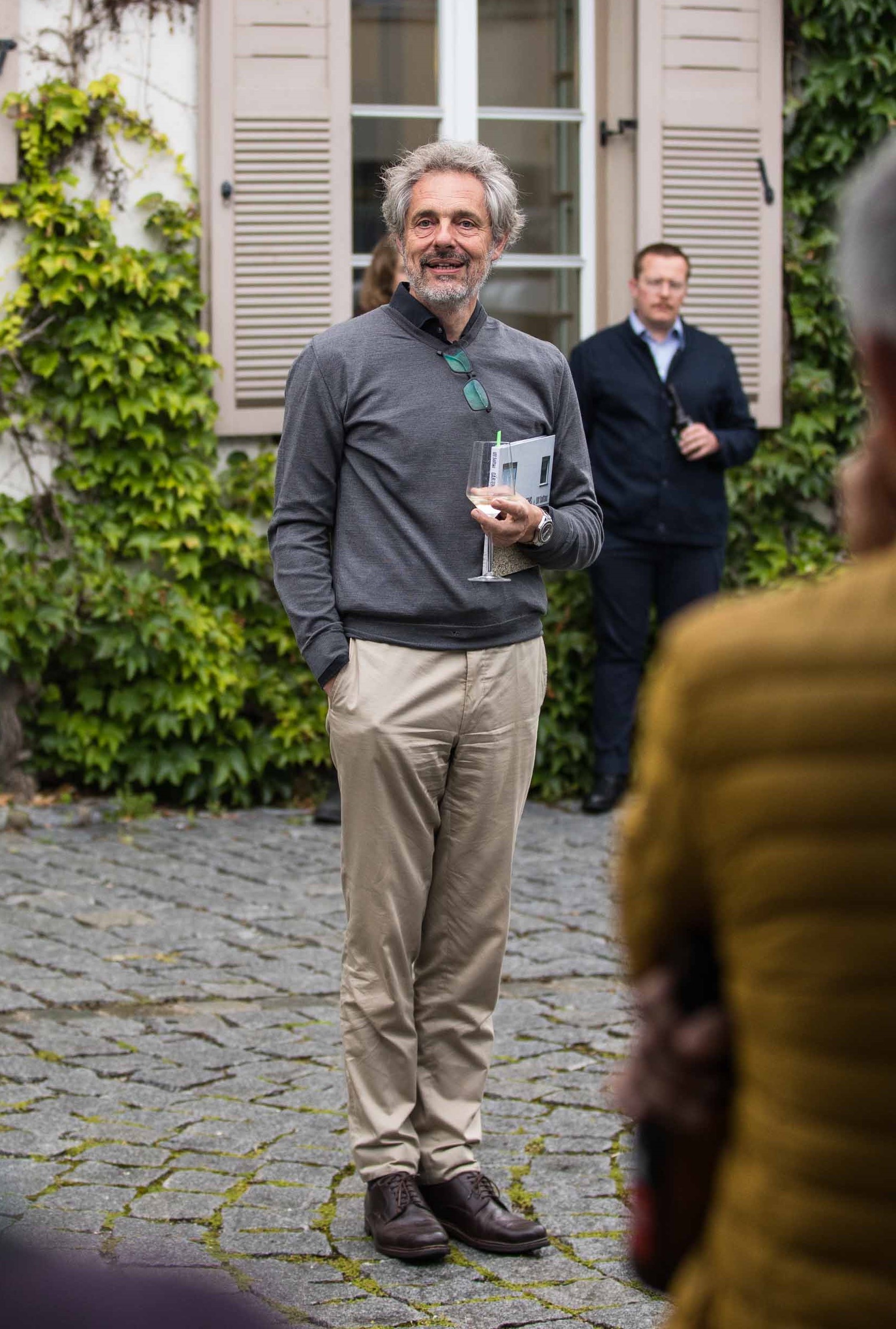 Ein Mann steht lächelnd in einem Innenhof vor einer klassischen Fassade mit Fensterläden und grünem Efeu.