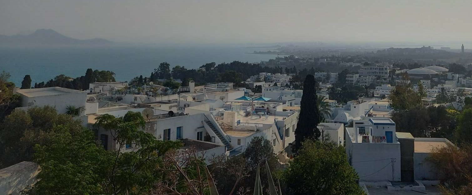 Weiße mediterrane Häuser mit flachen Dächern erstrecken sich über eine Küstenlandschaft mit Blick auf das Meer und entfernte Berge.