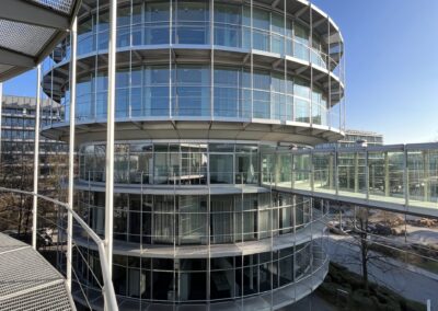 Ein modernes, zylindrisches Gebäude mit gläserner Fassade, weißen Stahlträgern und mehreren Etagen unter klarem Himmel.