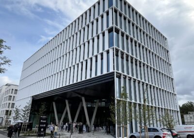 Ein modernes Hochhaus mit vertikaler weißer Lamellenfassade und großflächiger Glasfront im Erdgeschoss, umgeben von Fussgängern und Fahrzeugen.