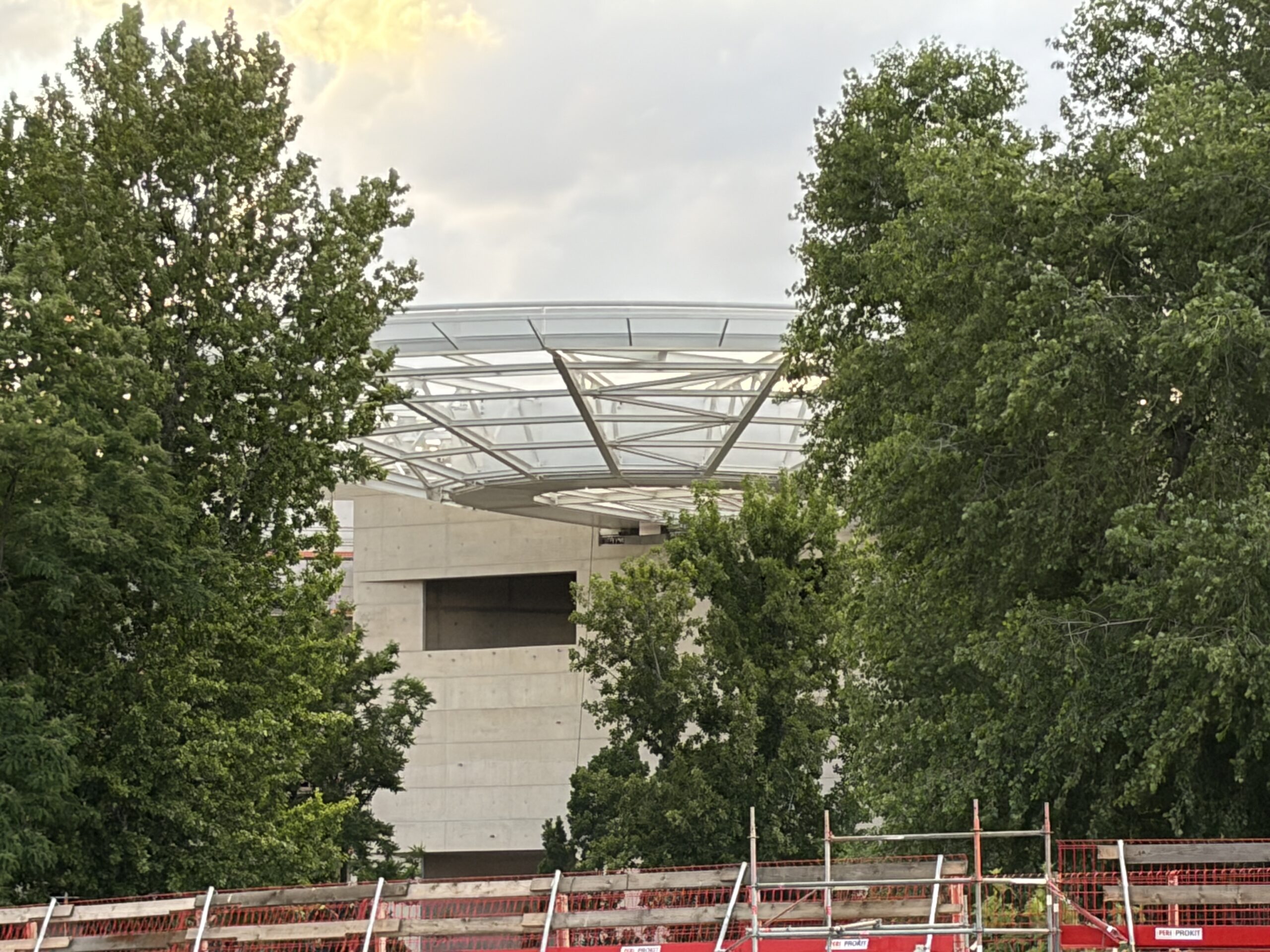 Zwischen Bäumen sichtbarer Sichtbetonbau mit weit auskragendem, filigranem Stahl-Glas-Dach über einer Baustelle.