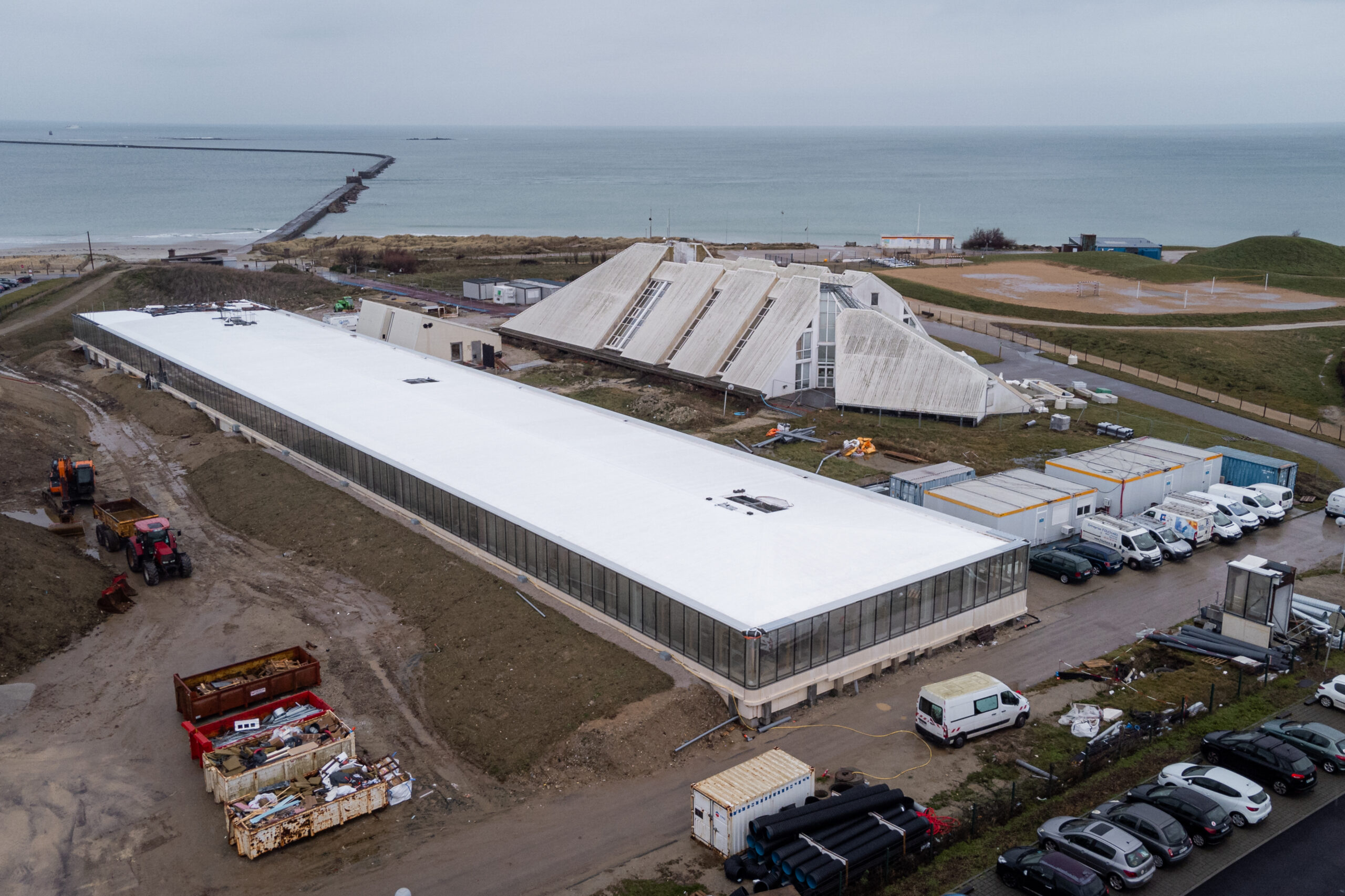 Langgestreckter Neubau mit weißem Flachdach und Glasfassade neben markanter Betonhalle mit gefalteten Dachschalen am Meer.