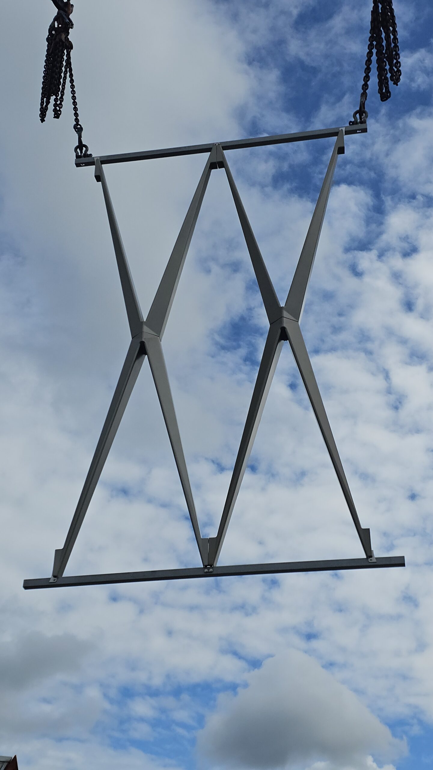 Ein an Kranketten hängendes Stahlfachwerk mit gekreuzten Diagonalstreben schwebt vor bewölktem blauem Himmel.